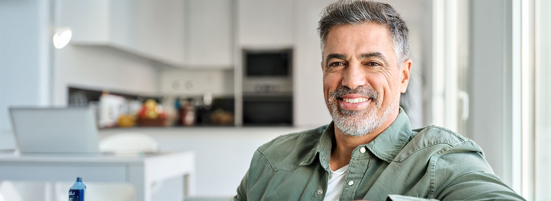 The image shows a man sitting at a kitchen counter with a smile on his face, wearing a green jacket and a white shirt, positioned in front of a window with blinds partially drawn.