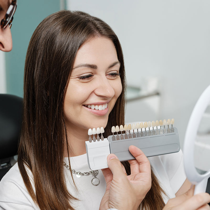 The image shows a woman sitting at a dental chair, holding a toothbrush with toothpaste, looking into a mirror, smiling, with her reflection visible on the mirror.