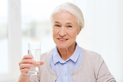 The image shows an elderly woman holding up a glass of water with her right hand while standing indoors.