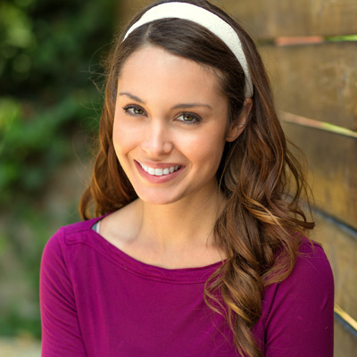 A woman with long brown hair, wearing a purple top and a white headband, poses for a portrait against a wooden fence backdrop.