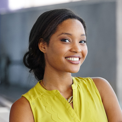 The image shows a smiling woman with dark hair wearing a yellow top, posing for a portrait against a blurred background.