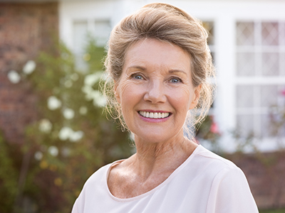 The image shows a woman with short hair smiling at the camera, wearing a white top, standing outdoors in front of a house with a brick facade.