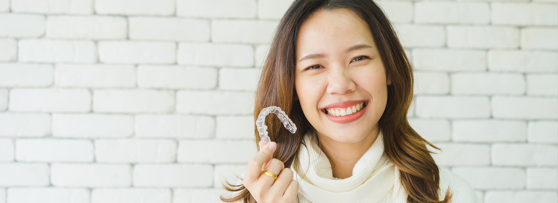 A smiling woman with dark hair holding a toothbrush, set against a white brick wall background.