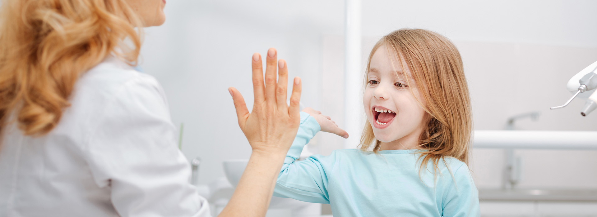 The image shows a young girl with her hands raised near her face, possibly clapping or waving, standing next to an adult woman who appears to be a mother, both indoors, with the child s reflection visible in a mirror behind them.