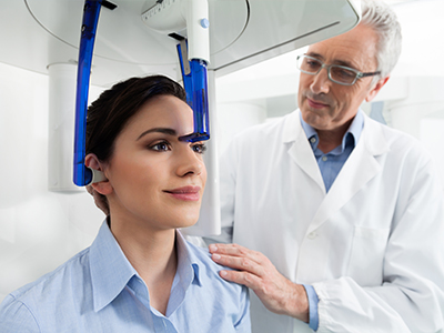 The image depicts a scene where a woman is seated in a dental chair with a large, blue, head-mounted device attached to her forehead, while a dentist stands behind her, observing the equipment and possibly explaining its use.