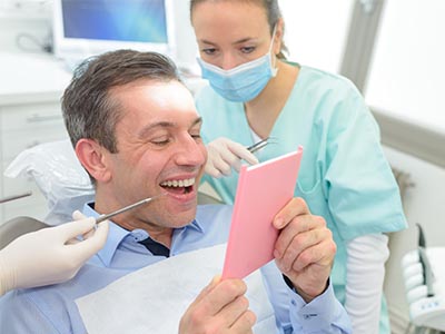 The image features a man sitting in a dental chair with his mouth open, looking at a pink card held by a woman wearing medical scrubs and a surgical mask, who appears to be showing him the card. There are two identical images side by side, creating a mirror effect.