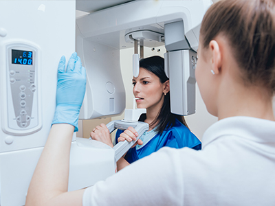 The image shows two individuals in a medical setting, with one person standing behind a large, advanced piece of diagnostic equipment, possibly an MRI machine, while another person stands in front, seemingly observing or preparing for the procedure.