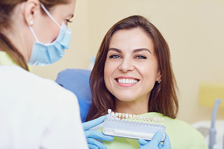 A woman with a bright smile is seated at a dental chair, receiving dental care from a professional who stands behind her, holding a dental tool.