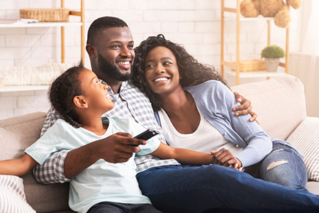 A family of four sitting together on a couch with a warm embrace, smiling at the camera.