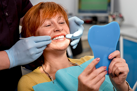 The image shows a woman sitting in a dental chair with a dental hygienist working on her teeth, smiling at the camera while holding up a blue toothbrush.