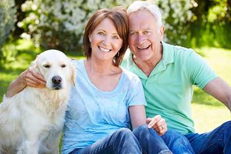 The image shows an older couple sitting outdoors with their golden retriever dog, both smiling and looking at the camera.