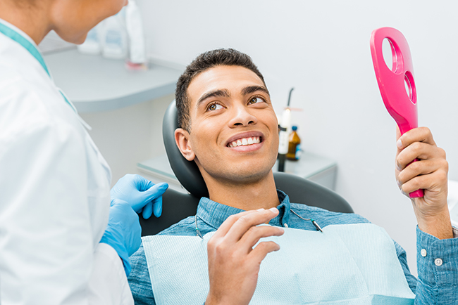 A man sitting in a dental chair with a smiling expression, holding a pink object that appears to be an oral care device, while surrounded by dental professionals in their work environment.