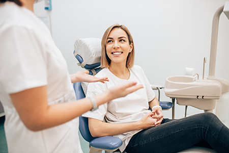 The image shows a woman sitting in a dental chair with her legs crossed, smiling at someone outside the frame, being attended to by two dental professionals who are focused on their work.