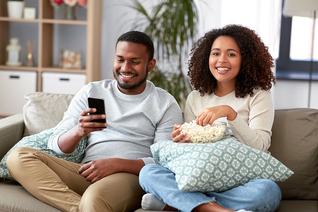 A man and woman sitting on a couch, sharing a moment with popcorn while watching something together, both smiling and engaged with each other.