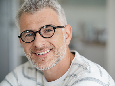 The image shows a man with glasses, smiling at the camera, against a blurred background.
