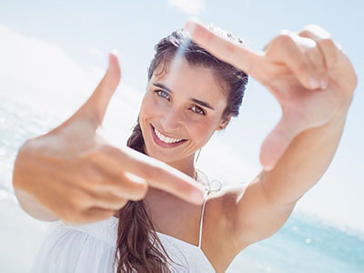 A young woman with light skin, long brown hair, and blue eyes smiles broadly while holding up two fingers against a bright background at the beach.