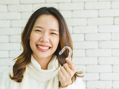 The image shows a smiling woman holding up a toothbrush with her right hand while standing indoors against a brick wall backdrop, next to a person whose face is partially obscured by a white object in their left hand.